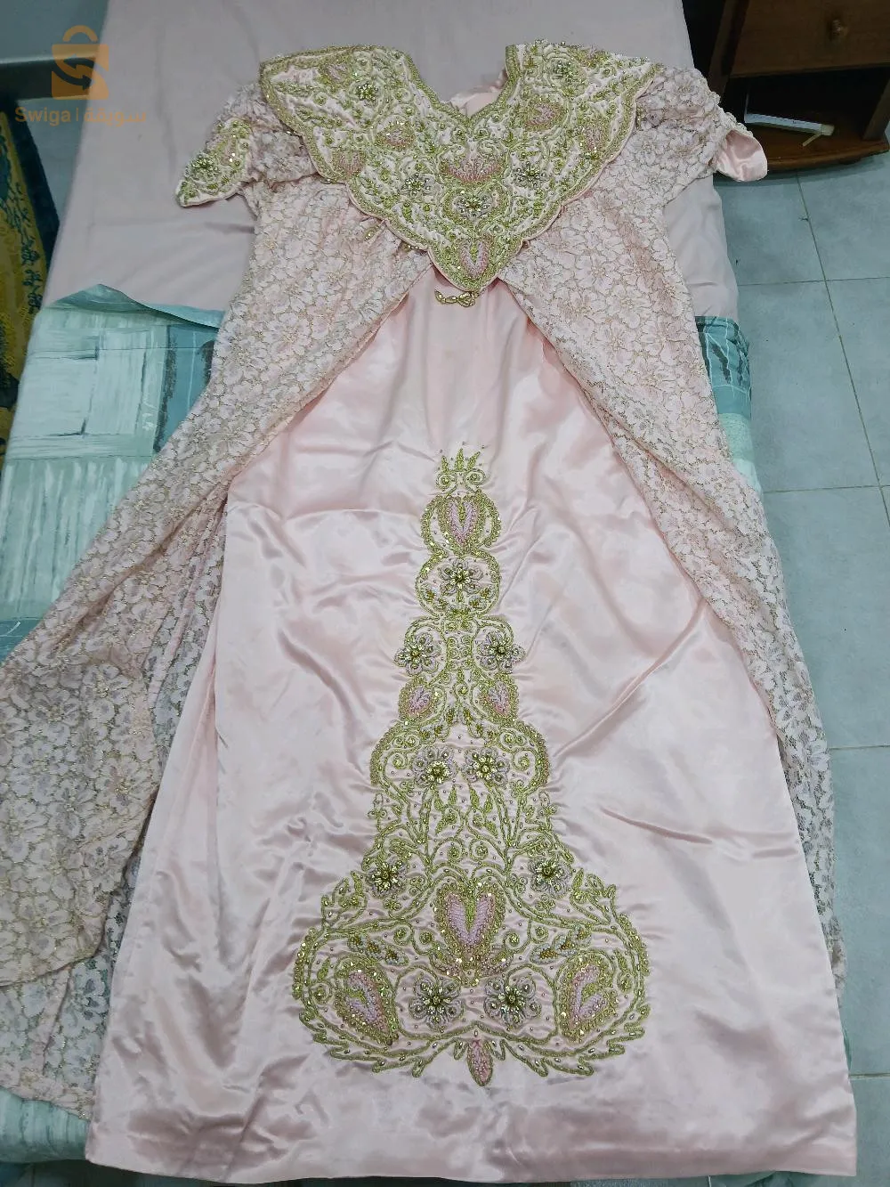 Cette robe Jeba Wahrani est une pièce traditionnelle algérienne élégante et raffinée, parfaite pour les grandes occasions. Confectionnée dans un tissu rose satiné, elle est ornée de superbes broderies en fil doré et perles qui ajoutent une touche de luxe et de glamour. Le col est richement décoré de motifs floraux et de détails minutieux, tandis que la jupe est agrémentée d'un grand motif central qui attire le regard. Les manches et les côtés de la robe sont accompagnés d'un voile en dentelle assorti, ce qui ajoute du volume et de l'élégance à l'ensemble. Cette robe Jeba Wahrani est idéale pour les mariages, les soirées et les événements traditionnels, offrant un style unique et distingué. En excellent état, elle est prête à sublimer votre garde-robe lors de vos occasions les plus spéciales.