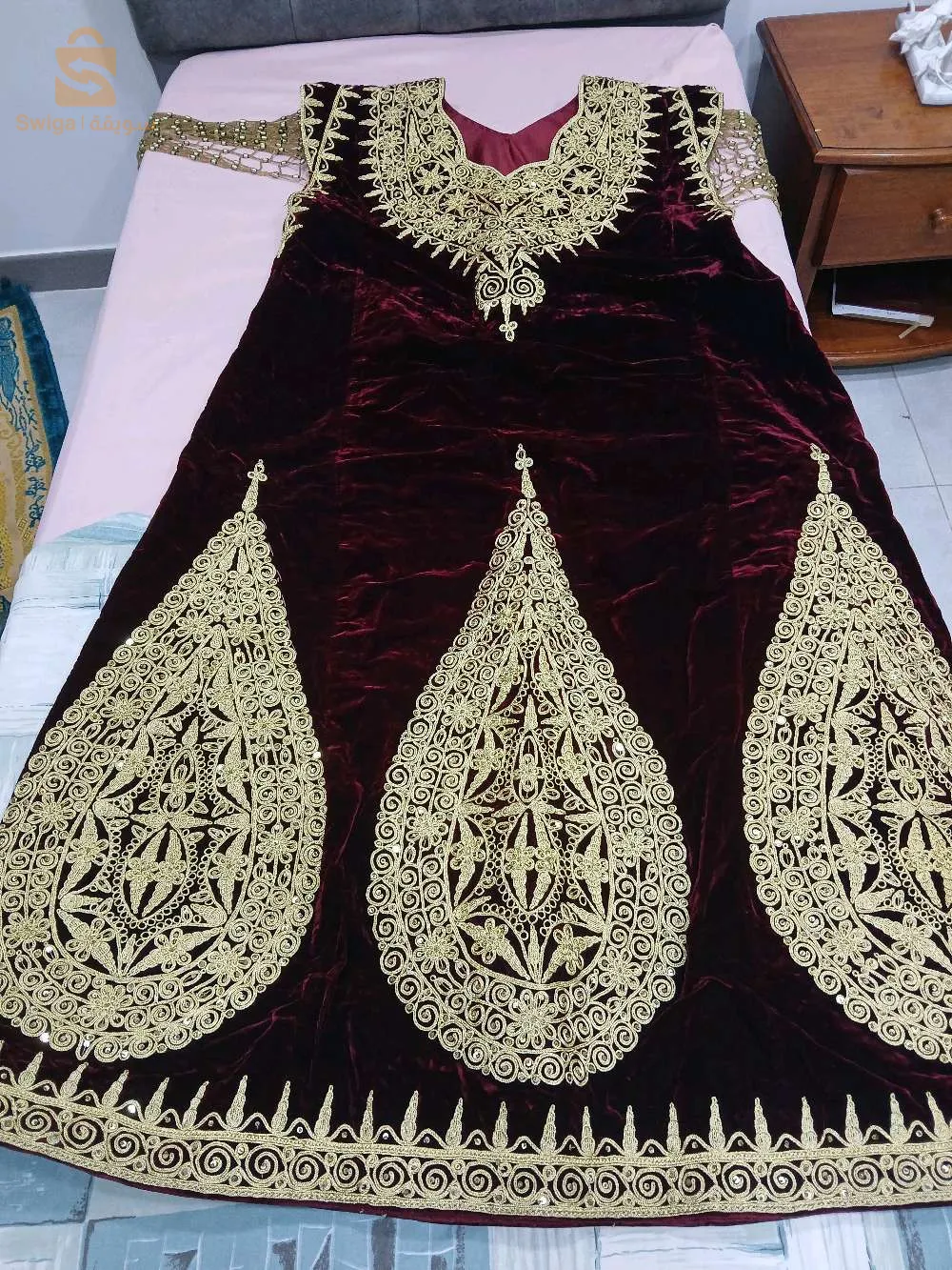 Cette robe Fergani est un magnifique exemple de l'artisanat traditionnel algérien. Confectionnée dans un tissu en velours bordeaux, elle est richement ornée de broderies en fil doré réalisées à la main, ce qui lui confère une apparence luxueuse et élégante. Le motif doré couvre l'ensemble de la robe, avec des détails complexes qui ajoutent une touche de sophistication. Le col est orné de broderies élaborées, tandis que le bas de la robe présente de grands motifs en forme de gouttes, un élément caractéristique du style Fergani. Cette pièce est idéale pour les occasions spéciales telles que les mariages, les fêtes ou les cérémonies traditionnelles. En excellent état et réalisée avec soin, elle représente une véritable œuvre d'art qui allie élégance et culture.