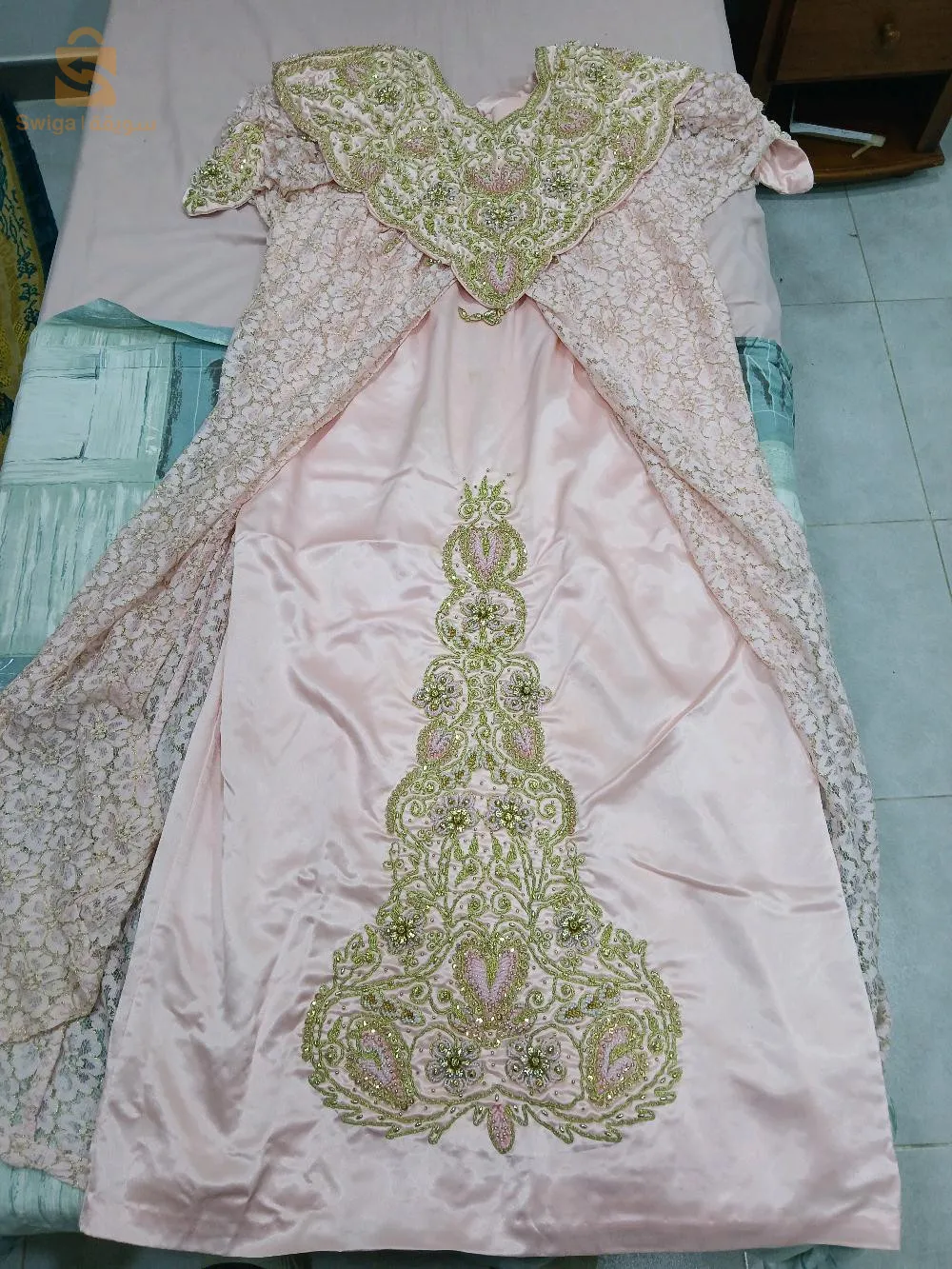 Cette robe Jeba Wahrani est une pièce traditionnelle algérienne élégante et raffinée, parfaite pour les grandes occasions. Confectionnée dans un tissu rose satiné, elle est ornée de superbes broderies en fil doré et perles qui ajoutent une touche de luxe et de glamour. Le col est richement décoré de motifs floraux et de détails minutieux, tandis que la jupe est agrémentée d'un grand motif central qui attire le regard. Les manches et les côtés de la robe sont accompagnés d'un voile en dentelle assorti, ce qui ajoute du volume et de l'élégance à l'ensemble. Cette robe Jeba Wahrani est idéale pour les mariages, les soirées et les événements traditionnels, offrant un style unique et distingué. En excellent état, elle est prête à sublimer votre garde-robe lors de vos occasions les plus spéciales.
