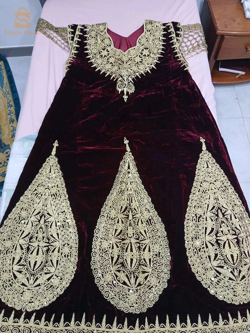 Cette robe Fergani est un magnifique exemple de l'artisanat traditionnel algérien. Confectionnée dans un tissu en velours bordeaux, elle est richement ornée de broderies en fil doré réalisées à la main, ce qui lui confère une apparence luxueuse et élégante. Le motif doré couvre l'ensemble de la robe, avec des détails complexes qui ajoutent une touche de sophistication. Le col est orné de broderies élaborées, tandis que le bas de la robe présente de grands motifs en forme de gouttes, un élément caractéristique du style Fergani. Cette pièce est idéale pour les occasions spéciales telles que les mariages, les fêtes ou les cérémonies traditionnelles. En excellent état et réalisée avec soin, elle représente une véritable œuvre d'art qui allie élégance et culture.